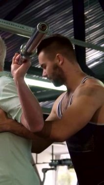İlham verici kıdemli bir adam spor salonunda halterle kendinden emin bir şekilde çömelir, dayanıklılık ve dayanıklılığı somutlaştırır. Kendini adamış genç bir eğitmen arkasında duruyor, sarsılmaz bir destek ve rehberlik sunuyor,