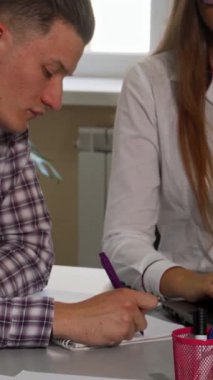 Kızıl saçlı bir kadın ve kısa saçlı bir adam sınıf masasında yoğun bir şekilde birlikte çalışıyorlar, ifadeleri kararlılık ve konsantrasyonu yansıtıyor. Enerjik atmosfer akademik kovalamacanın enerjisiyle dolu.