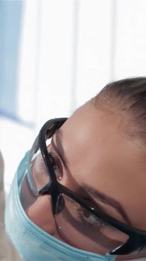 Gıcır gıcır beyaz üniformalı genç, sarışın bir dişçinin yakın çekimi ışıl ışıl bir gülümsemeyle muayeneyi bitiriyor. Nazikçe koruyucu gözlüklerini ve maskesini çıkarıyor, diş hekimleriyle dolu, parlak, klinik bir ortamda sıcaklık ve profesyonellik yayıyor.