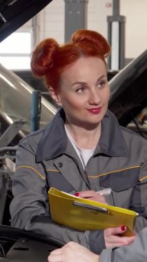 Canlı kızıl saçlı yetenekli bir bayan tamirci ve erkek arkadaşı otomotiv dünyasında özenle çalışan bir araba motorunu inceliyor. Şık gri üniformalı kadın, parlak sarı bir panoya titiz notlar alır.