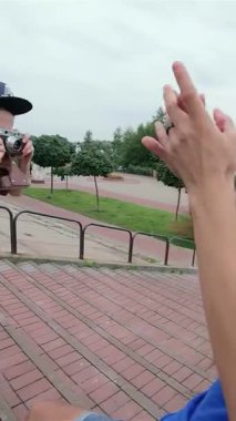 Genç bir adam neşeli bir şekilde gülen arkadaşlarının açık havada, yemyeşil ve bulutlu bir gökyüzüyle çevrili canlı bir merdivende fotoğraflarını çeker. Bu canlı sahne arkadaşlığın özünü yansıtıyor, kahkaha ve aşkla dolu tasasız yaz günlerini yakalıyor.