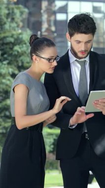 Dinamik bir iş çifti, çağdaş mimari ve verimli yeşilliklerle süslenmiş şık bir şehir ofisinde tablet üzerinde işbirliği yaparak başarılarından zevk alıyor. Onların kendine güvenen gülümsemeleri ve şık profesyonel kıyafetleri hırs ve takım çalışması yayıyor.