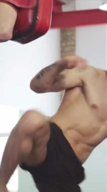 Büyüleyici düşük açılı bir atış, güçlü bir tekme atan kaslı sakallı bir erkek dövüşçüyü sergiliyor, koçu ise ustalıkla tekme pedallarını tutuyor. Enerjik spor salonu ayarları enerji ve yoğunluk yayar, dövüşçülerin adanmışlığını vurgular ve