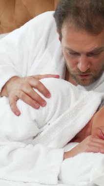 Sıcak, aydınlık bir yatak odasında, olgun bir kadın yatakta kıvrılıp yatar, yüzü hayal kırıklığını yansıtırken, kocası yumuşak beyaz bir bornoz giyer ve konuşmak için eğilir. Bu samimi ama gergin an, ilişkilerinin duygusal mücadelesini yakalar.