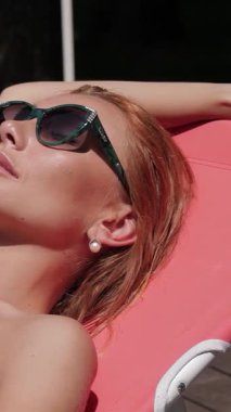 Göz kamaştırıcı kızıl saçlı bir kadın zarif bir iskemlede güneşlenir, parlak bir yüzme havuzunun yanında güneşlenir. Canlı pembe bir bikini ve şık bir güneş gözlüğü takmış yaz cazibesi ve kaygısız bir mutluluk yayıyor.
