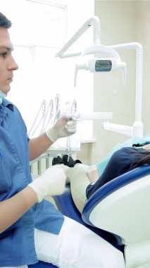 Gösterişli, modern bir dişçi kliniğinde, mavi üniformalı genç bir erkek dişçi ileri ışıklandırma ekipmanları kullanarak kadın hastaların dişlerini dikkatlice inceliyor. Bu samimi sahne diş hijyeninde hassasiyeti ve özeni vurguluyor.