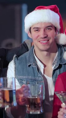 Canlı bir üçlü, iki erkek ve bir kadın neşeli Noel Baba şapkaları giyer ve neşeli bir barda kadehlerini kaldırırken neşeli bir şekilde kadeh kaldırırlar. Atmosfer kahkaha ve tatil neşesi ile uğulduyor, renkli dekorasyonlar ve sıcak