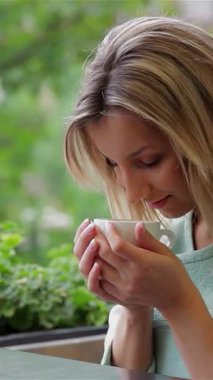 Genç bir iş kadını, dalgalı sarı saçları olan, dumanı tüten bir fincan kahveyi nazikçe kucaklıyor, onun zengin aromasını sakin bir gülümsemeyle soluyor. Rahat bir kafede yemyeşil bir zemin üzerine kurulmuş bu an rahatlığı ve neşeyi güzelce özetliyor.