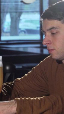 Neşeli genç bir adam, elinde soğuk bir bardak zanaat birası, sıcak, davetkar bir barın ışıltısını tutarken neşe saçıyor. Yumuşak akşam ışığı sahnede dans eder. Rahatlamış atmosferi canlandırır. Rahatlamak ve zevk almak için mükemmeldir.