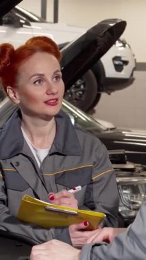 Canlı kırmızı bukleleri olan çekici bir kadın tamirci, modern arabalarla dolu hareketli bir garaja notlar yazarak araç tamirleri hakkında canlı bir tartışmaya girişiyor. Bu dinamik sahne onun uzmanlığını ve özgüvenini ön plana çıkarıyor.