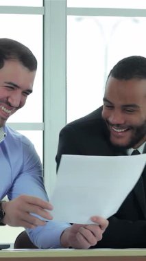 Biri açık mavi gömlekli, diğeri parlak siyah takım elbiseli iki kendine güvenen işadamı, güneşli bir ofiste önemli belgeleri incelerken başarılarını sevinçle kutluyorlar. Parlak gülümsemeleri ve çekici ifadeleri güçlü bir şeyi yansıtır.