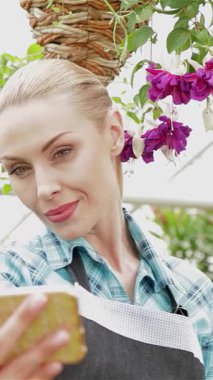 Işıl ışıl beyaz bir çiçekçi, şen çiçeklerle ve şen yeşilliklerle çevrili, yemyeşil bir serada neşeli bir selfie çekerken ışıl ışıl gülümsüyor. Şık bir önlük ve ekose gömlek giyen bu çekici sarışın kadın, neşeyi temsil ediyor.