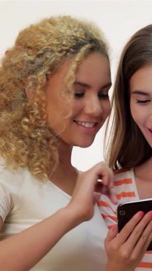 İki kadın, biri kıvırcık kıvırcık sarı saçlı, diğeri uzun kahverengi saçlı, tablet bir bilgisayarla heyecan verici bir şekilde etkileşime geçiyor, beyaz bir zemine neşe saçıyorlar. Parlak gülümsemeleri ve günlük kıyafetleri enerjik bir hava yaratıyor.