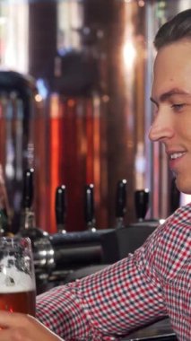 Şık, mutlu bir adam canlı bir bar ortamında arkadaşıyla canlı bir sohbete dalmış taze bir biranın tadını çıkarır. Barın sıcacık parıltısı ve davetkar dekor atmosferi güçlendiriyor. Bu da onu dostluk ve neşe dolu mükemmel bir an yapıyor.