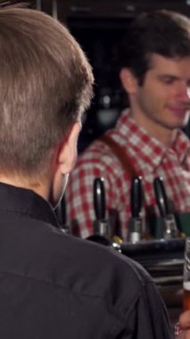 Şık, mutlu bir adam soğuk birasının tadını çıkarıyor, canlı bir bar ortamında bir arkadaşıyla sohbet ediyor. Sıcak ışıklandırma ve kırsal dekor, dostluğu ve güzel zamanları kutlamak için mükemmel davetkar bir atmosfer yaratır.