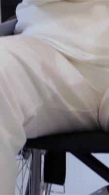 Üst düzey bir beyaz kadın parlak, modern bir rehabilitasyon odasında etrafındakileri yansıtırken düşünceli bakışları aşağıya doğru çevrilmiş parlak bir tekerlekli sandalyede oturuyor. Yumuşak, ılık ışık, onun nazik özelliklerini vurgulayarak, bir huzur yaratıyor.