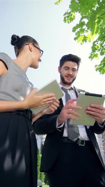 Kendine güvenen bir işadamı ve profesyonel bir iş kadını parlak mavi gökyüzünün sıcaklığında güneşlenerek tabletlerinde fikirlerini paylaşıyorlar. Işıl ışıl gülümsemeleri ve çekici ifadeleri arka planda yer alan başarılı bir ortaklığı yansıtıyor.