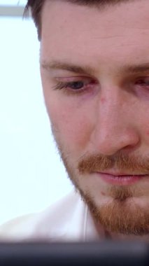 Kararlı genç bir beyaz erkek yönetici öne eğiliyor, delici mavi gözleri modern ofis pencerelerinden akan parlak doğal ışıkla aydınlanan dizüstü bilgisayara kilitlenmiş. Şık beyaz gömleği ve bakımlı sakalı yansıtıyor.