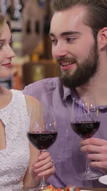 Neşeli genç bir çift, sıcak, mum ışığında bir restoranda gözleri aşkla parıldayan kırmızı şarap kadehlerini paylaşır. Zarif tabaklarla çevrili ve sıcak bir ortam ile, yakın ilişkileri mutluluk yayıyor.