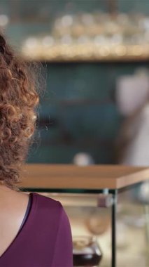 Kıvırcık saçlı neşeli genç bir kadın, sıcak, davetkar bir kafede kahvesinin tadını çıkarırken mutluluk saçıyor. Dost canlısı bir barista ustalıkla arkasından içki hazırlıyor. Lezzetli atmosfer, taze ve baştan çıkarıcı aromasıyla zenginleşir.