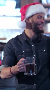 Şenlikli bir barda, neşeli Noel Baba şapkaları takan iki arkadaş buzlu bira bardaklarını kaldırarak bayram ruhunu somutlaştırıyorlar. Neşeli atmosfer, gülümseyen bir barmen ve parıldayan ışıkların sıcacık parıltısı ile güçlenir.