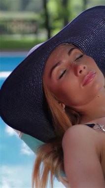 Göz kamaştırıcı beyaz bir kadın sıcak güneşin altında lüks içinde, koyu mavi bir mayoyla ışıldayan bir havuzun kenarında uzanır. Onun uzun, dalgalı sarı saçları ve şık geniş kenarlı şapkası yaz mutluluğunun bir resmini oluşturuyor, rahatlama hissi uyandırıyor ve...