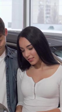 Neşeli bir çift sıcak bir kucaklaşmayı paylaşıyor, parlak yeni araba anahtarlarını lüks araçlarla dolu, modern bir galeride gururla sergiliyorlar. Güzel kadın heyecanla ilk otomobillerini satın almalarının kilometre taşını kutluyor.