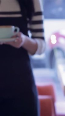 Şık çizgili gömlekli ve önlüklü neşeli bir bayan garsonun yakın çekimi pastel mavi bir fincanda nefis bir kahve servisi yapar, sıcaklık ve konukseverlik yayar. Parlak gülüşü ve özenli tavırları davetkar bir atmosfer yaratıyor.