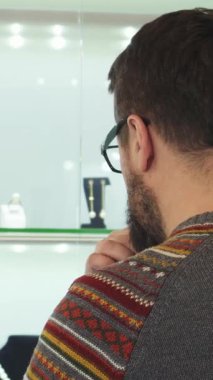 Sade, desenli süveterli sakallı bir adam, göz kamaştırıcı bir mücevher sergisine derinden odaklanmış, hareketli bir alışveriş merkezinde duruyor. Düşünceli tavrı, yürekten bir hediye seçmenin heyecanını yakalıyor. Havada romantizm ve zerafet yayıyor.