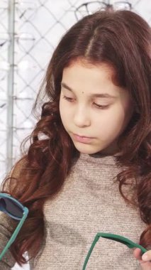 Uzun, dalgalı kahverengi saçlı hoş bir kız elinde iki tane şık güneş gözlüğü olan canlı bir gözlük mağazasında duruyor. Yüzü kafa karışıklığı ve merakı yansıtıyor. Renkli gözlüklerden oluşan bir kaleydoskop ile çevrelenmiş,