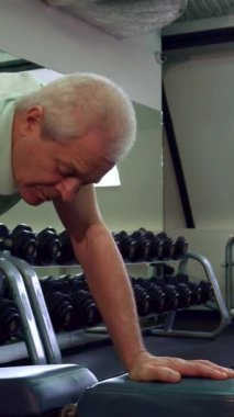 Yaşlı beyaz bir adam spor salonundaki bir dambılı kaldırırken, kaşları konsantrasyondan çatılmış bir şekilde kararlılık sergiliyor. Yakınlarda, genç bir antrenör teşvik edici bir atmosfer sunuyor, bu hareketli fitness alanı ile dolu