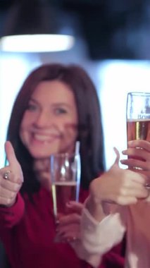 Genç yetişkinlerden oluşan canlı bir üçlü, iki kadın ve bir erkek, canlı bir barda arkadaşlığı kutlamak için soğuk bir bardak birayla coşkuyla tezahürat yapıyor. Işıltılı gülümsemeleri ve coşkulu jestleri arkadaşlığın heyecanını özetliyor.