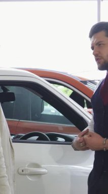 Özel dikim takım elbiseli parlak bir araba satıcısı zarif bir şekilde beyaz bir aracın kapısını açar. Kırmızı bir fiyonkla süslenir ve erkek müşterisini lüks içinde yaşamaya davet eder. Galerideki bu dinamik an heyecan yayıyor.