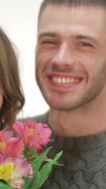 Neşeli bir çift sıcak bir kucaklaşmayı paylaşır. Yumuşak beyaz arka planda çarpıcı bir buket canlı çiçek tutarlar. Samimi bir kazak ve şenlikli bir Noel Baba şapkası giyip ışıl ışıl gülümsemeleri ve ışıl ışıl gözleriyle aşkın ve bayramın özünü yakalıyor.