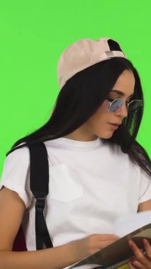 Şık bir beyzbol şapkası ve şık güneş gözlükleriyle süslenmiş ışıl ışıl genç bir kız öğrenci, canlı yeşil renkli bir arka plana karşı canlı kitaplarını gururla tutarken neşeyle ışıldıyor. Bu dinamik sahne gençliğin ruhunu özetliyor.