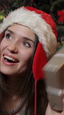 Neşeli esmer bir kadın, neşeli bir Noel Baba şapkası takıyor, güzel paketlenmiş bir Noel hediye kutusunu neşeyle sallıyor, yüzü saf zevk saçıyor. Işıl ışıl süslerle ve parıldayan ışıklarla süslenmiş ışıl ışıl bir Noel ağacıyla çevrili.