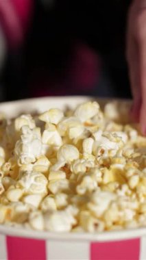 Tüylü, altın patlamış mısırla dolu canlı bir kova film gecesinin heyecanını yakalar. Oyuncak ojelerle süslenmiş bir kız eli, hevesle suya dalıyor, neşe saçıyor ve lezzetli sinemalarda atıştırmalıklar bekliyor.