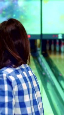 Şık mavi ve beyaz kareli gömlekli neşeli bir esmer kız bovling oynamaya hazırlanıyor, gözleri önlerindeki renkli iğneleri hedeflerken heyecandan parıldıyor. Canlı bowling salonu göz kamaştırıcı ışıklar ve canlı bir atmosferle dolu.