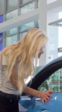 Modern bayilikte çarpıcı mavi bir arabayı özgüvenle incelerken, dalgalı sarı saçlı ışıl ışıl genç bir kadın heyecanla parıldıyor. Bu canlı sahne araba sahibi olmanın heyecanını, güzelliği ve çağdaş yaşam tarzını özetliyor.