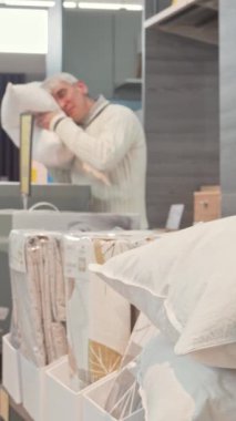 Gümüş saçlı yaşlı bir adam, bir dizi yumuşak tekstil ile çevrili şık bir mobilya mağazasında ortopedik yastığı dikkatle inceler. Modern dekorla vurgulanan davetkar atmosfer ve düzgünce düzenlenmiş yatak,