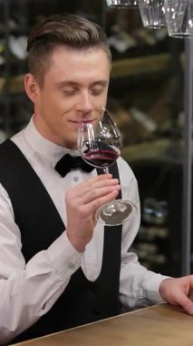 Özel dikim yelek ve papyon giymiş kendine güvenen bir şarap garsonu lüks bir kırmızı şarabın derin aromasının tadını çıkarır. Minnettarlık içinde gözleri kapalıdır. Enfes bir şarap rafının arka planına kurulmuş bu sahne sofistike bir şekilde ışıldıyor.