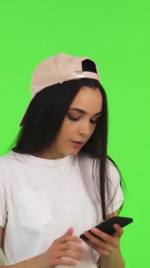 Işıl ışıl, siyah saçlı ve şık şapkalı genç bir kadın akıllı telefonuyla canlı yeşil bir krom arka planda selfie çekiyor. Bulaşıcı gülüşü ve şakacı tavırları modern sosyal medyanın özünü yansıtıyor.