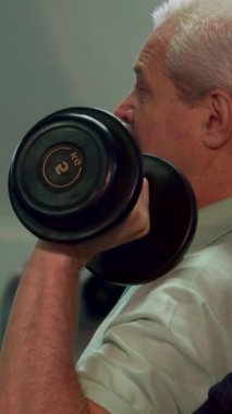 Kararlı, gücünü gösteren yaşlı bir adam, güç antrenmanı sırasında sarsılmadan bir dambılı kaldırır. İfadesindeki yoğunluğu yakin plan yaklasiyor. Formunu belirleyen adanmışlik ve direnci vurguluyor.