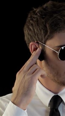 Havacı gözlüğü ve parlak siyah kravatı olan şık giyimli bir güvenlik görevlisi, çarpıcı bir siyah arka plana karşı etrafını yoğun bir dikkatle tarıyor. Dengeli hareketleri ve ciddi ifadesi otoriteyi ve hazırlığı simgeliyor.