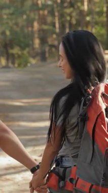 Neşeli bir çift yürüyüşçü, görkemli ağaçlar ve benekli güneş ışığıyla çevrili güneşli bir orman patikası boyunca el ele yürüyor. Çekici beyaz erkek ve kadın, açık hava maceralarını kucaklarken mutluluk saçıyor, aşkı somutlaştırıyor ve