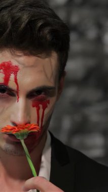 Gösterişli siyah takım elbiseli çarpıcı bir erkek vampir canlı turuncu bir çiçeği nazikçe burnuna kaldırarak kanla lekelenmiş yüzüne karşı rahatsız edici bir kontrast oluşturuyor. Koyu, desenli tuğla bir duvara karşı dizilmiş bu büyüleyici görüntü ürkünç görüntüyü harmanlıyor
