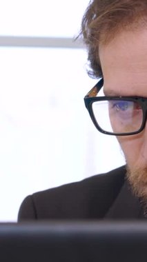 Sivri giyimli, titizlikle hazırlanmış sakallı beyaz bir işadamı yoğun bir şekilde dizüstü bilgisayarına odaklanmış ve mali veri denizine dalmış durumda. Pencereden gelen yumuşak arka ışık, onun ciddi yüz ifadesine sıcak bir ışık saçıyor.