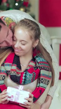 Sıcak, renkli kazaklara ve canlı eşarplara sarılı neşeli bir çift, güzel bir şekilde süslenmiş bir odada güzel bir hediyeyi tutarken şefkatli bir kucaklaşmayı paylaşıyor. Büyüleyici atmosfer, yanıp sönen ışıklar ve yemyeşil bir Noel ile aydınlanıyor.