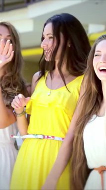 Canlı yaz elbiseleri giymiş üç modaya uygun kız renkli alışveriş çantalarını tutarken coşkuyla el sallıyor, neşe ve dostluk saçıyorlar. Işıl ışıl gülüşleri ve kaygısız kahkahaları bulaşıcı bir atmosfer yaratıyor.