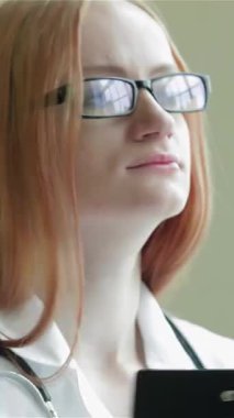 Kendini işine adamış kızıl saçlı ve şık gözlüklü genç bir kadın hasta ile ilgilenmeye hazır bir pano elinde duruyor. Pencereden süzülen yumuşak, sıcak ışık profesyonelliğini ve merhametini arttırır.